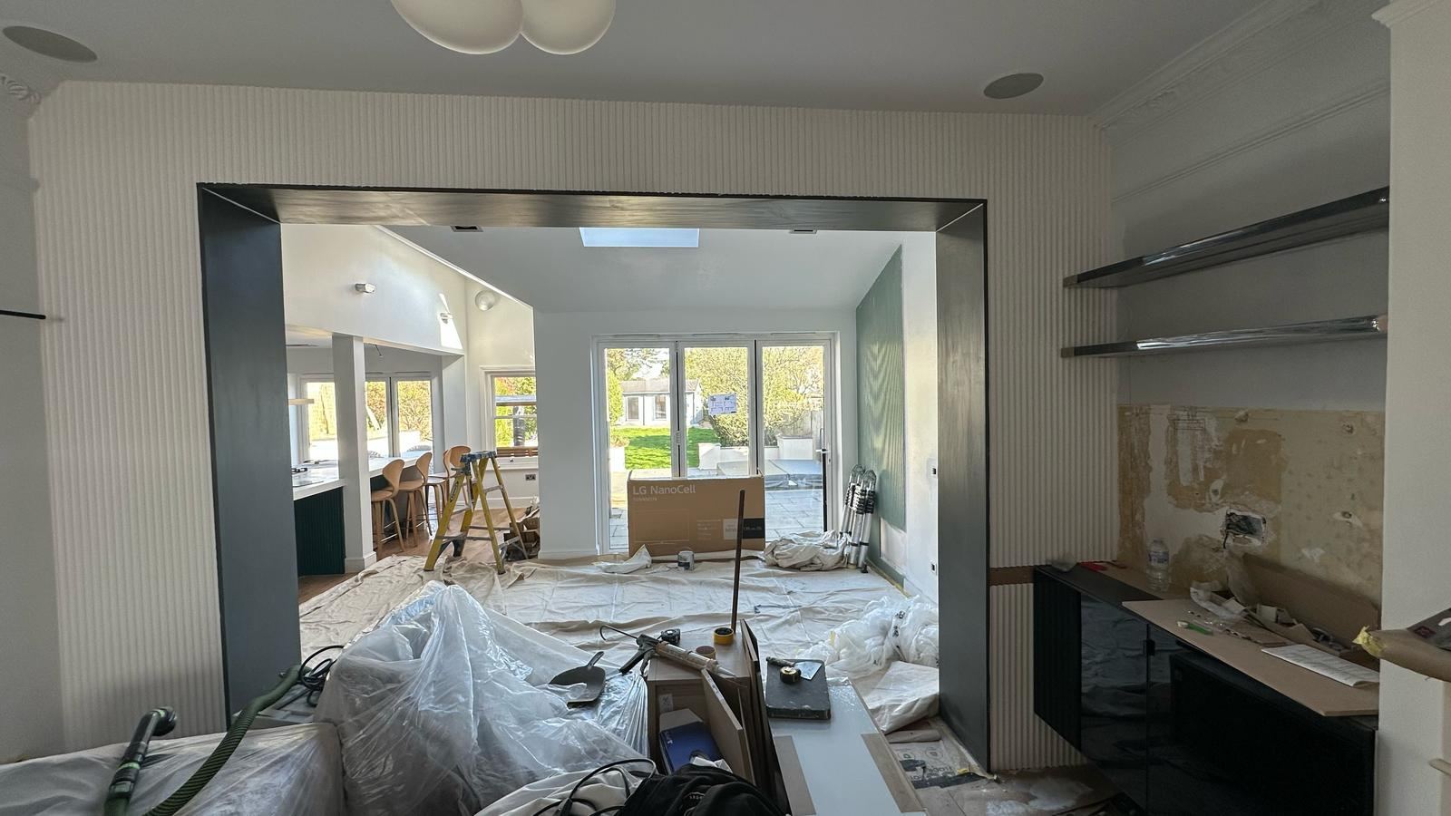 The dark-lined opening between the front room and the kitchen during the work, the floor sheeted and the far doors open to the garden